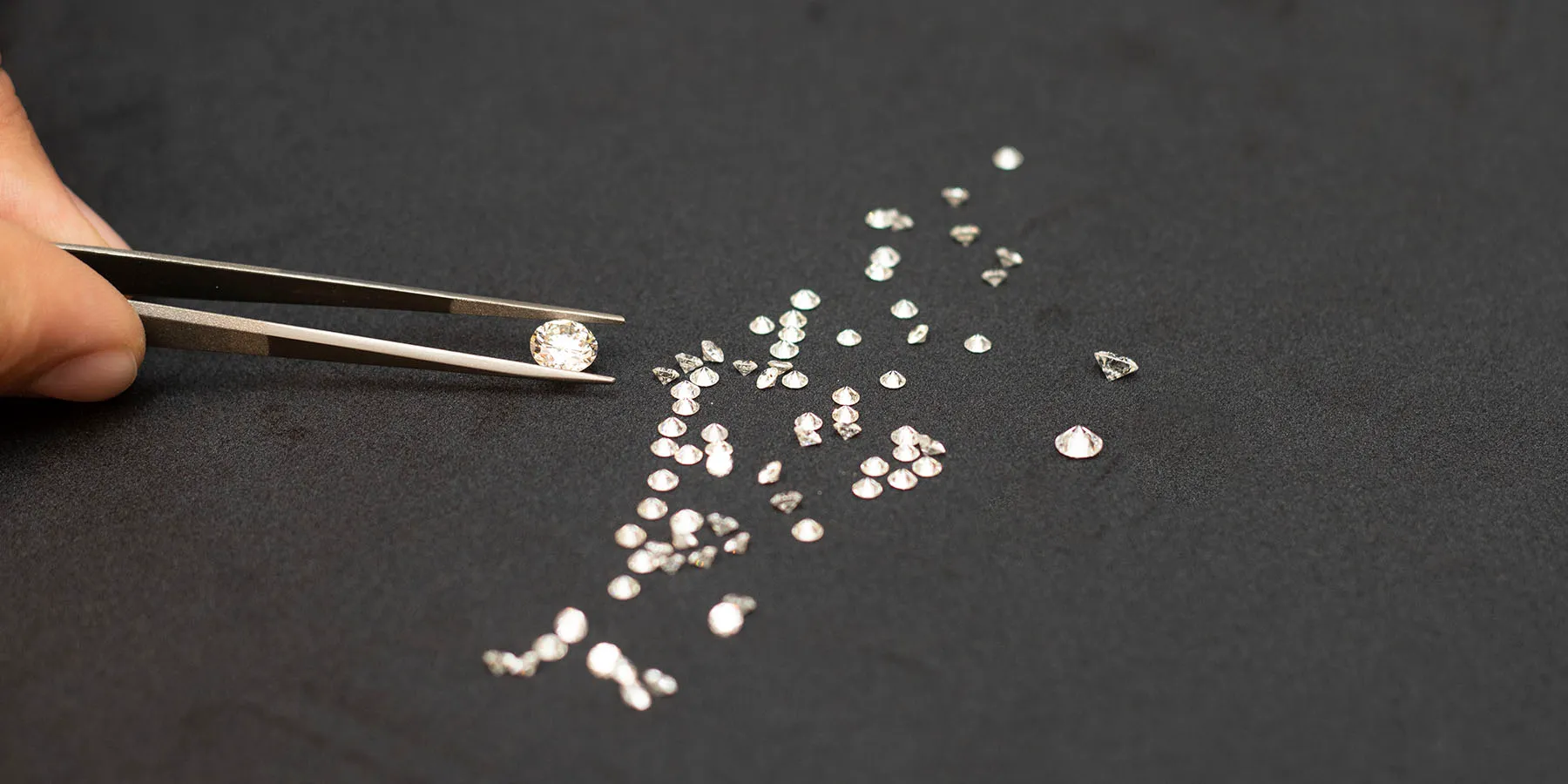 Tweezers holding a diamond above loose diamonds on a black cloth